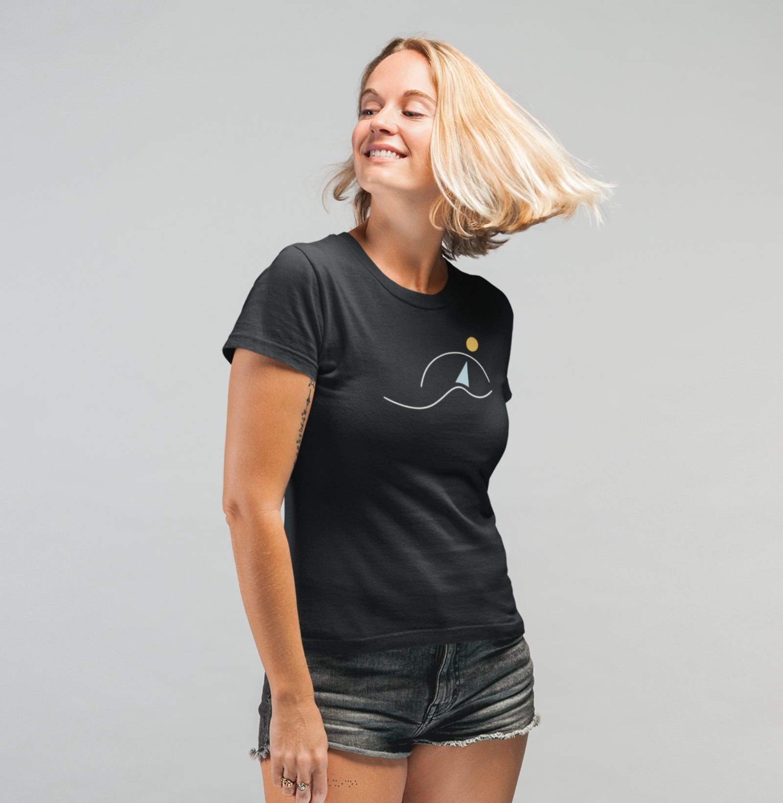 Mulher sorrindo usando camiseta preta À Beira do Dia da Latitude23, com estampa de veleiro minimalista sob o sol, refletindo liberdade e elegância náutica.