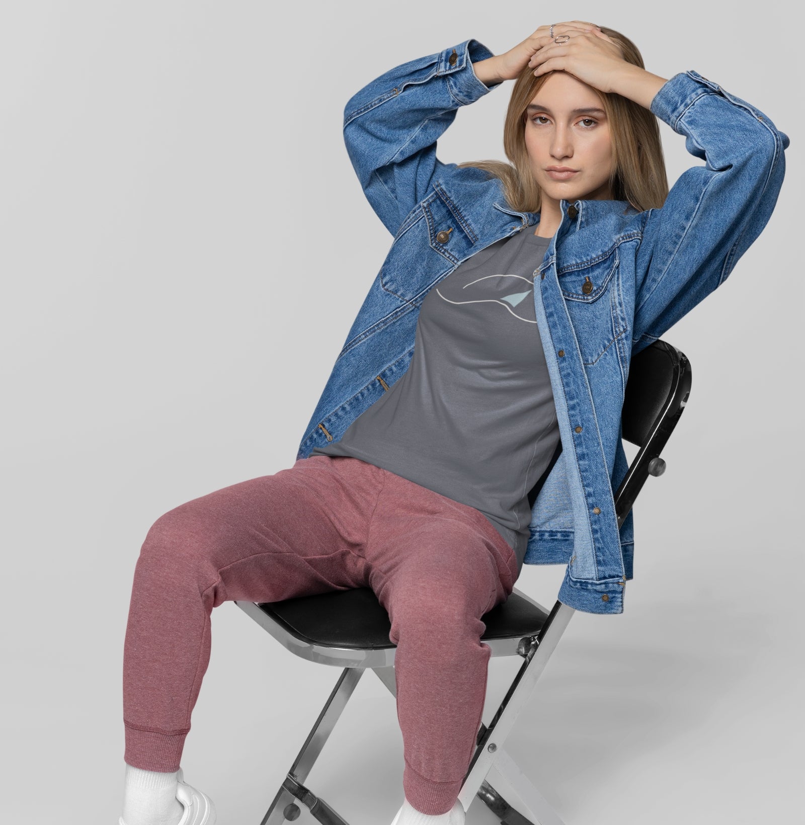 Mulher sentada com jaqueta jeans aberta usando camiseta cinza À Beira do Dia da Latitude23, exibindo estampa minimalista inspirada no horizonte marítimo.