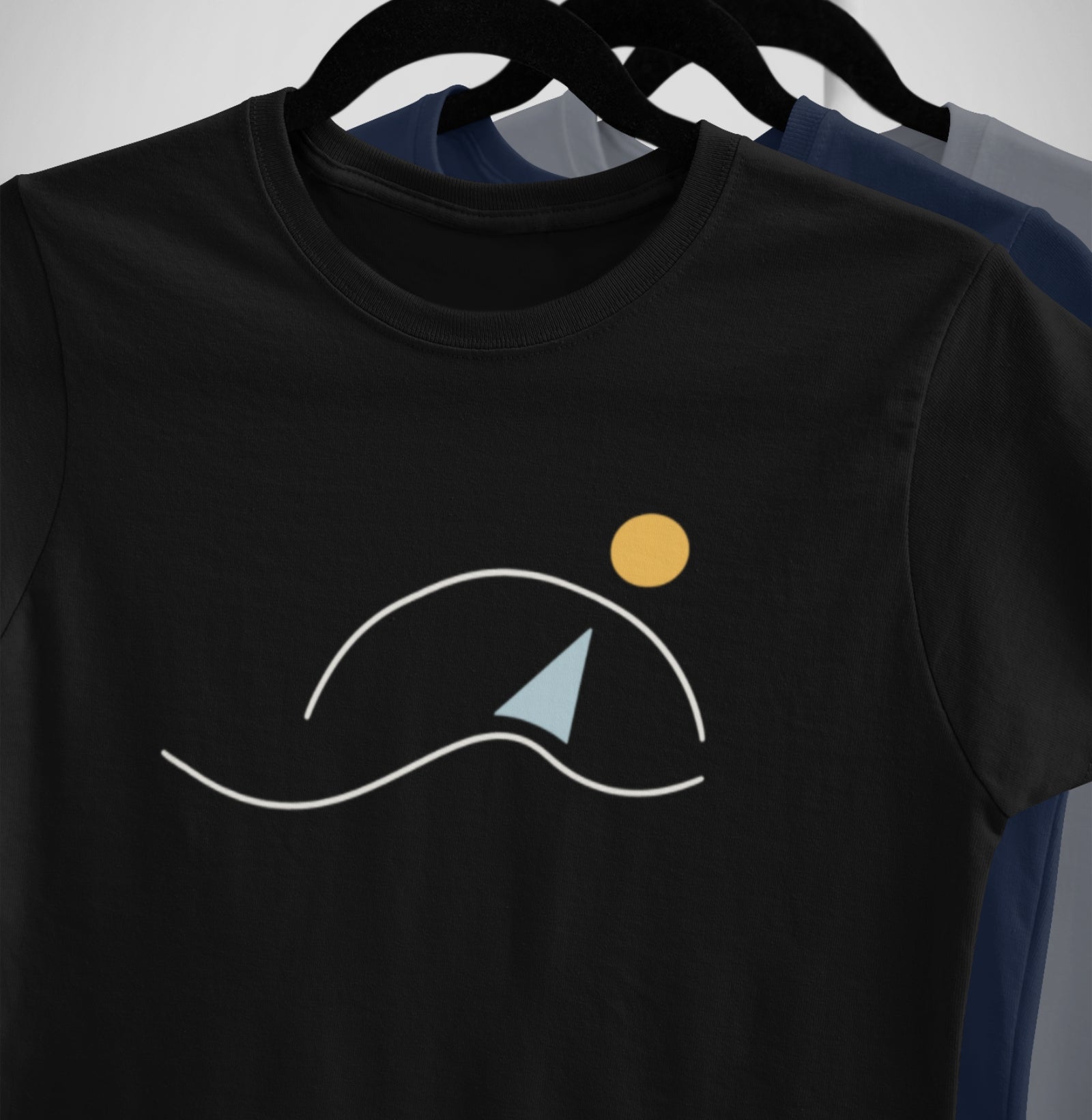 Camisetas À Beira do Dia da Latitude23 penduradas em cabides, exibindo estampa minimalista de veleiro e sol, símbolo de calmaria e elegância náutica.