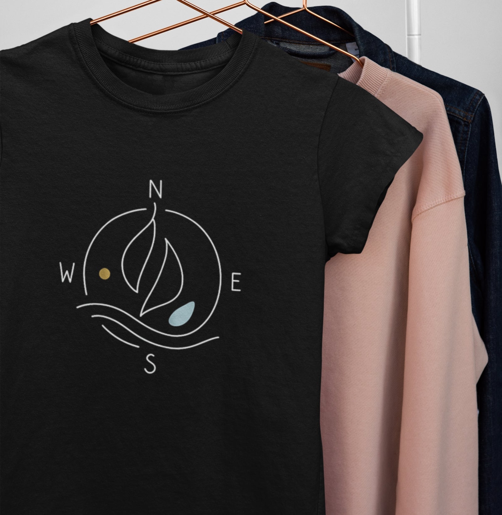 Camiseta preta Between Four Winds da Latitude23 pendurada em cabide, exibindo estampa náutica minimalista com bússola e vela, símbolo de liberdade e direção.
