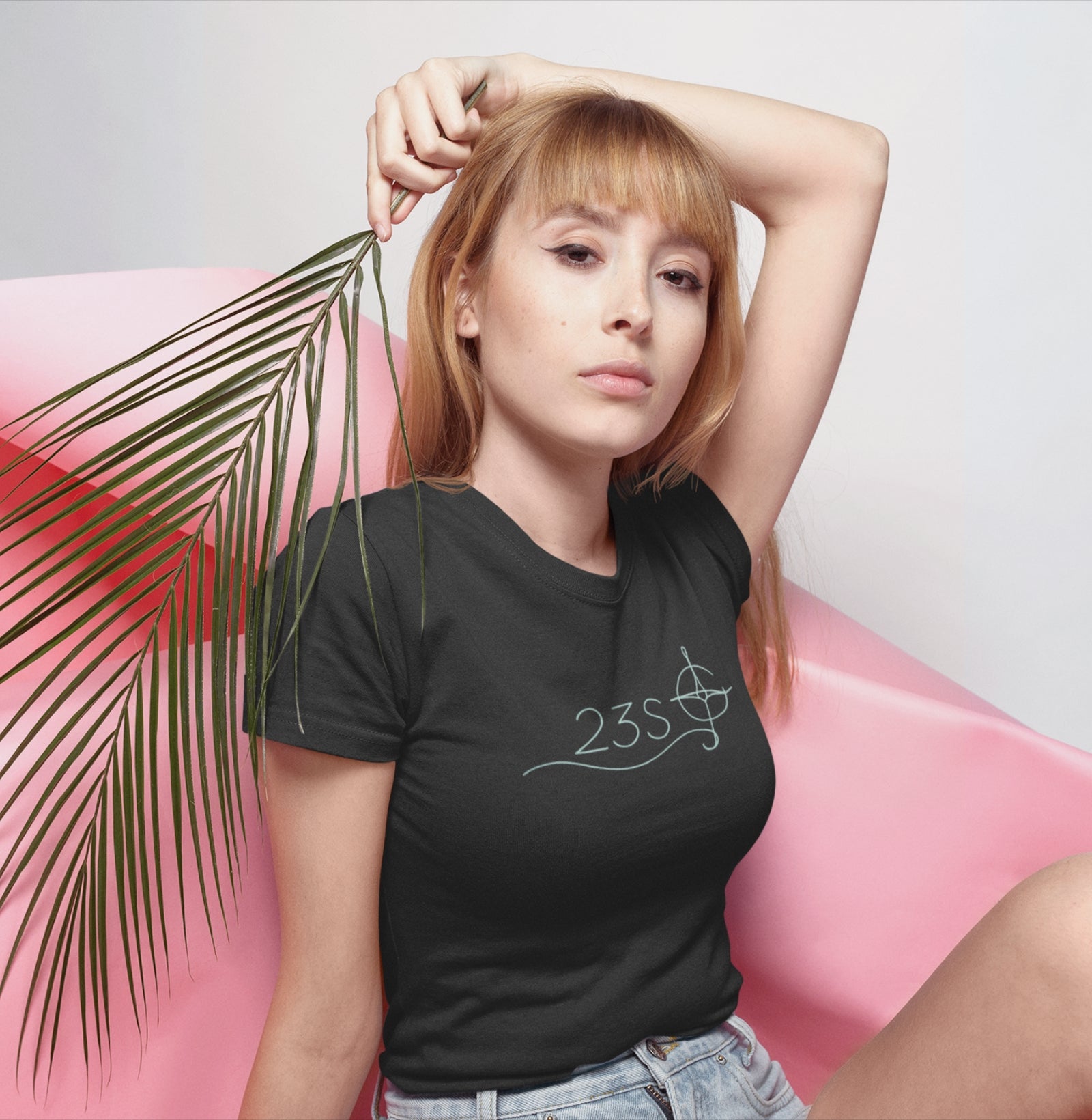 Camiseta preta Blue Axis da Latitude23 com design 23S e símbolo de bússola, usada por mulher loira em pose elegante com fundo rosa e folha tropical.