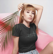 Camiseta preta Blue Axis da Latitude23 com design 23S e símbolo de bússola, usada por mulher loira em pose elegante com fundo rosa e folha tropical.