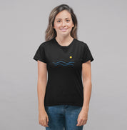 Camiseta preta Blue South da Latitude23 com design minimalista de ondas azuis e sol dourado, usada por mulher sorridente com jeans em fundo neutro.