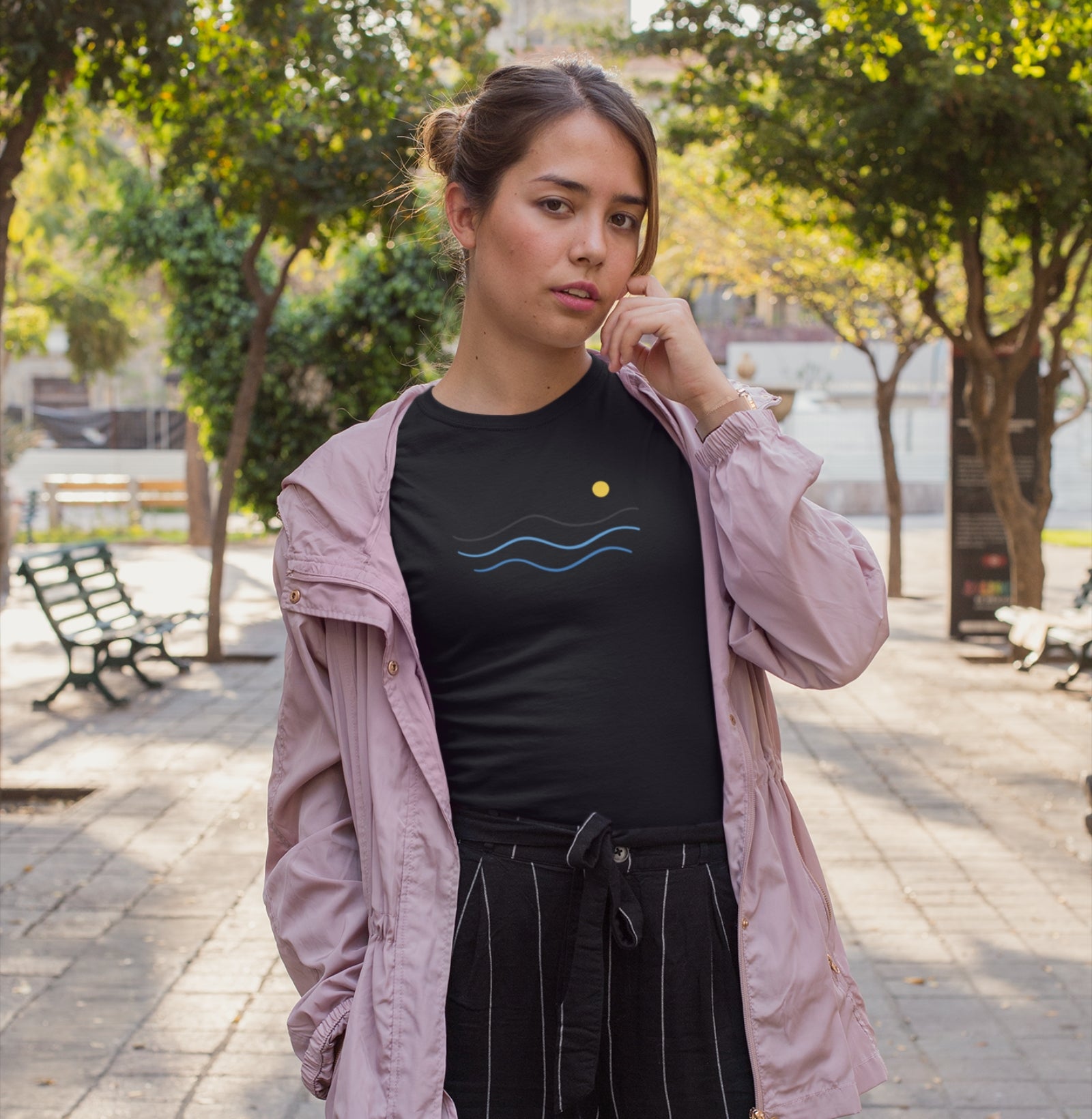 Camiseta preta Blue South da Latitude23 com design minimalista de ondas azuis e sol dourado, usada por mulher em ambiente urbano com jaqueta rosa clara.