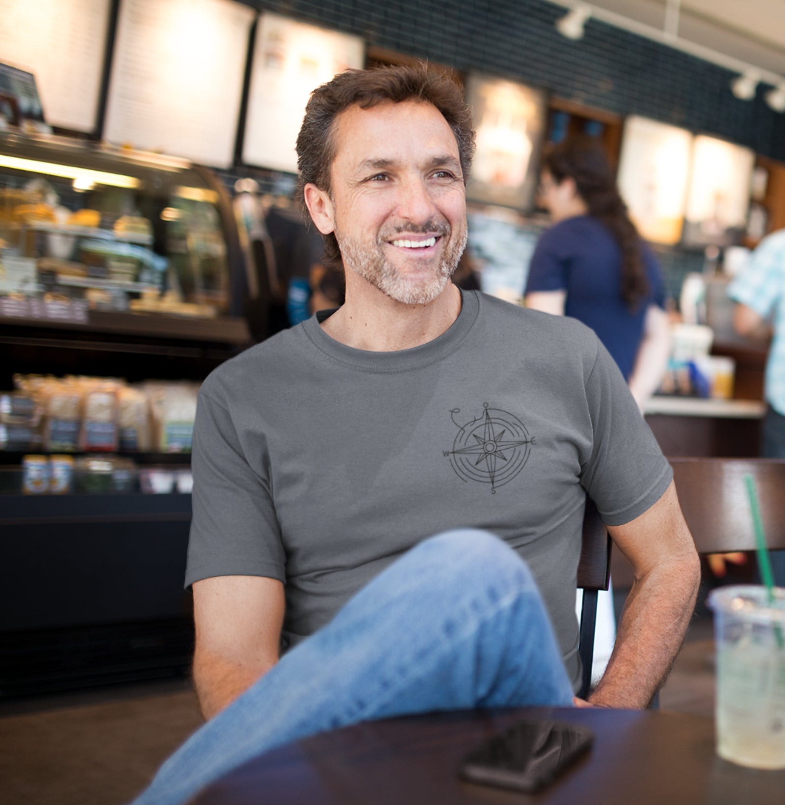 Homem usando a camiseta cinza Guiding Wind da Latitude23 em um café, mostrando a estampa de rosa dos ventos e o estilo náutico casual e sofisticado da marca.