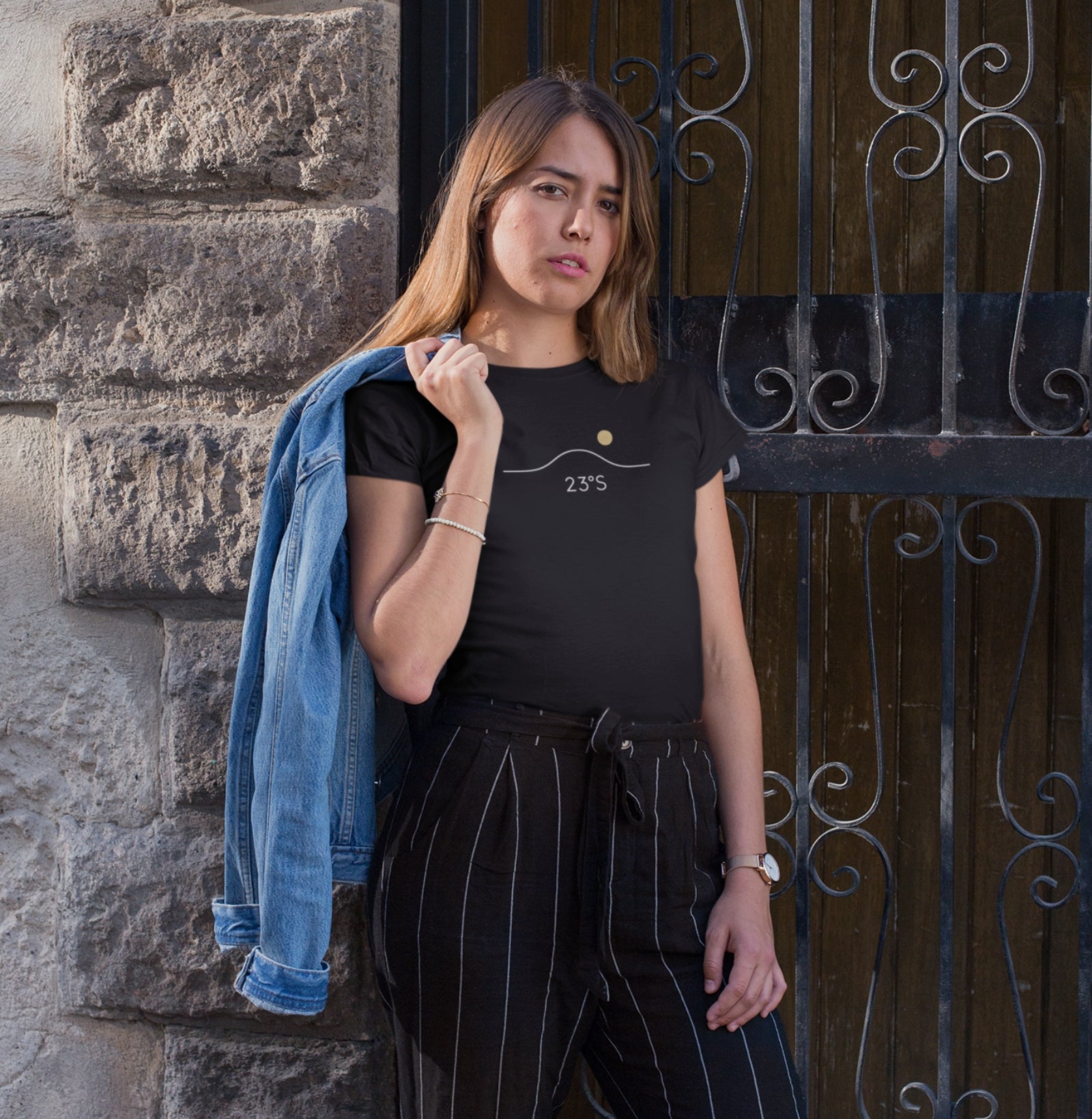 Camiseta preta Horizon 23°S da Latitude23 com design minimalista do horizonte e sol dourado, usada por mulher com jaqueta jeans no ombro em ambiente urbano.