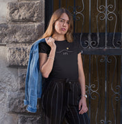 Camiseta preta Horizon 23°S da Latitude23 com design minimalista do horizonte e sol dourado, usada por mulher com jaqueta jeans no ombro em ambiente urbano.