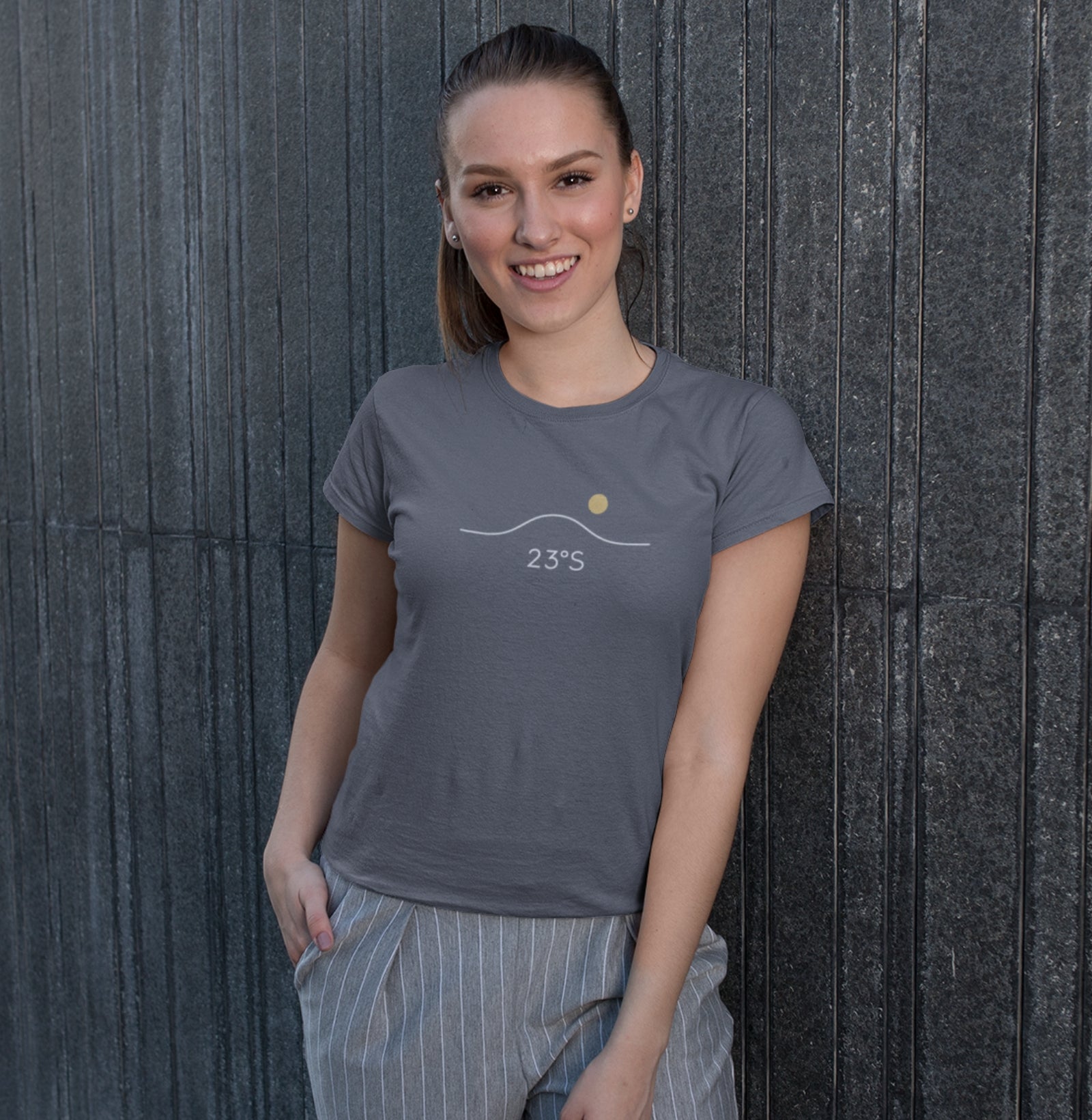 Camiseta cinza Horizon 23°S da Latitude23 com design minimalista de horizonte e sol dourado, usada por mulher sorridente com calça listrada em parede urbana escura.