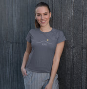 Camiseta cinza Horizon 23°S da Latitude23 com design minimalista de horizonte e sol dourado, usada por mulher sorridente com calça listrada em parede urbana escura.