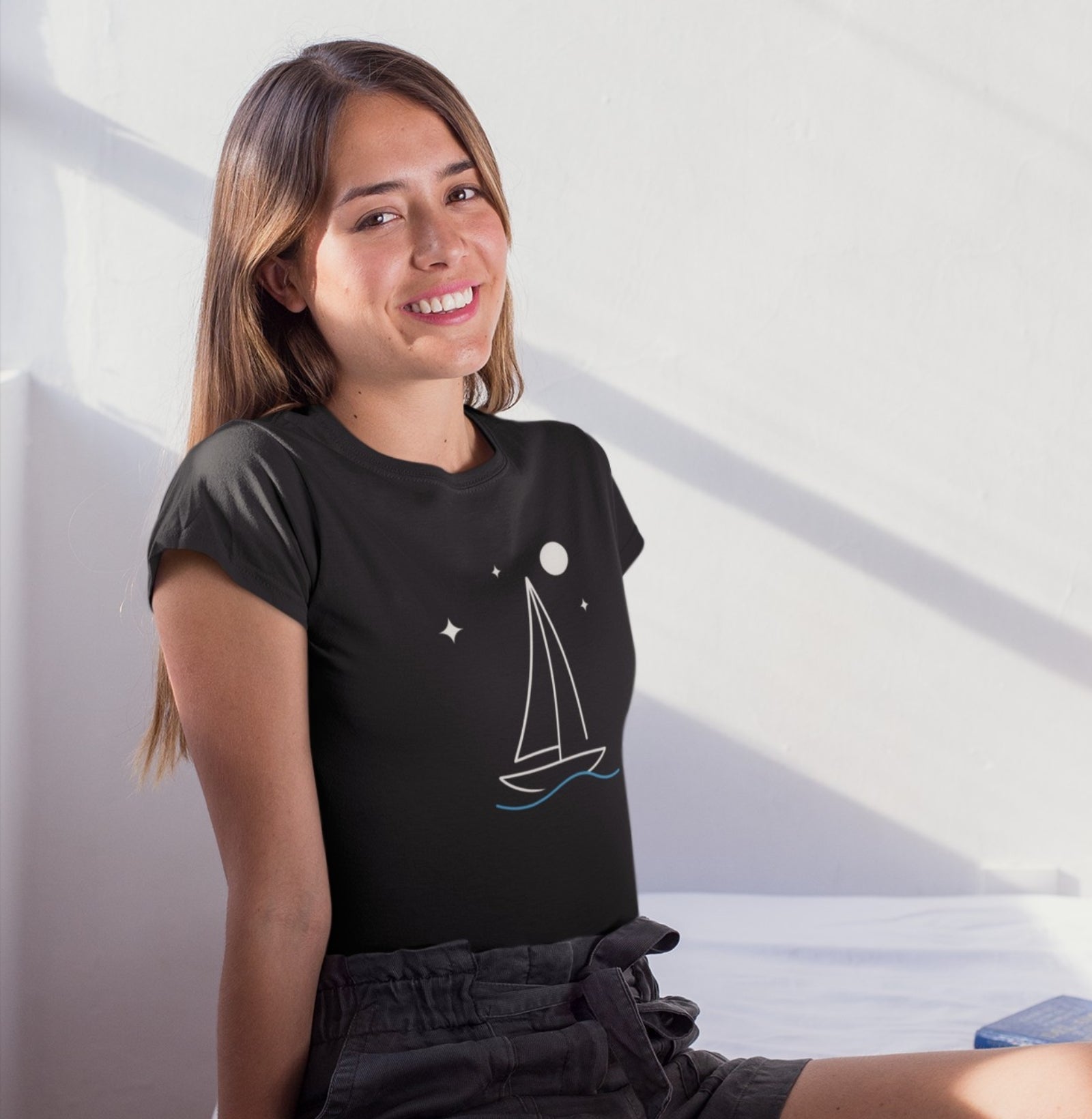 Mulher sorrindo com camiseta preta Horizonte de Prata da Latitude23, exibindo estampa minimalista de veleiro sob estrelas e lua, inspirada na calma do mar noturno.